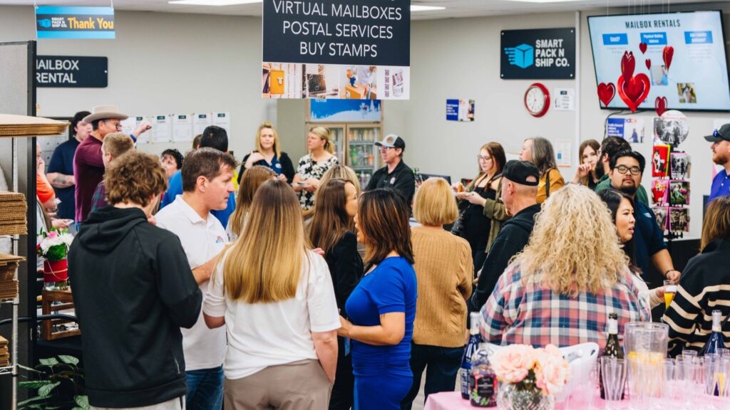 Busy retail store interior with customers mingling, signs for 'Virtual Mailboxes' and 'Postal Services', reflecting a successful opening.