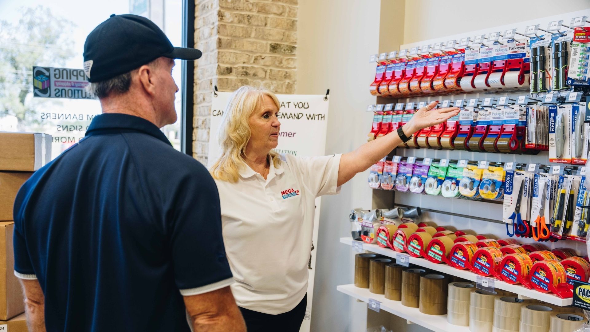 Mega Store Builders consultant in a "Mega Store Builders" polo shirt pointing to a display of shipping supplies while talking to a client.